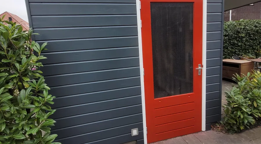 De geschilderde schuur over de volle breedte: donkergrijze delen, een oranjerode deur met wit kozijn en een laurierhaag ernaast