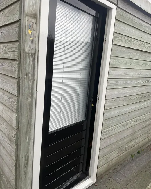 Een verweerde grijsgroene houten gevel met een donkere deur met lamellen en een raam met jaloezieën