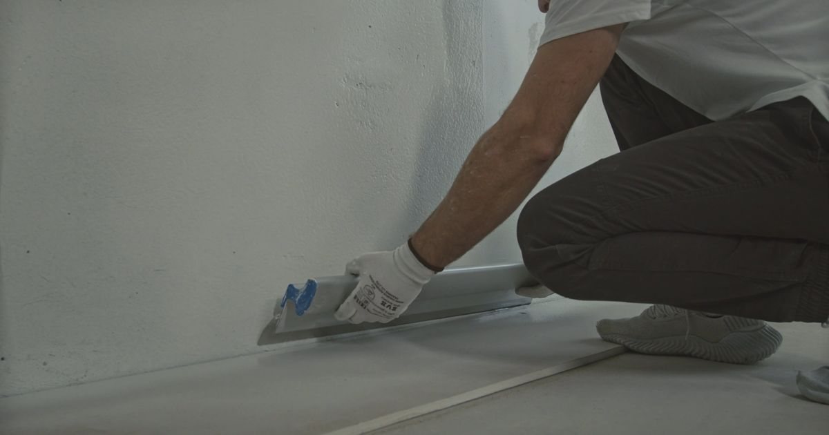 Een stukadoor op één knie trekt een brede stalen spaan langs de onderkant van een witte wand