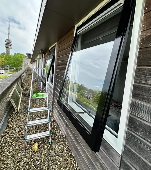 Een houten gevel met een groot nieuw raam, een aluminium trap ervoor en grind langs de gevel