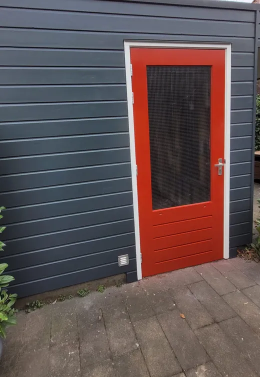 Dezelfde schuur vanaf dezelfde hoek, met strak geschilderde donkergrijze delen en een oranjerode deur