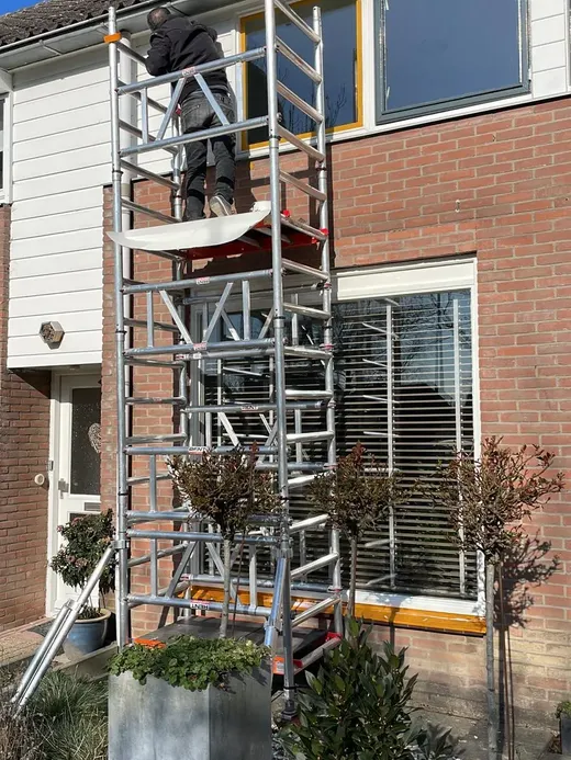 Een aluminium rolsteiger tegen een woning van baksteen en wit stucwerk, met bovenin iemand aan het werk