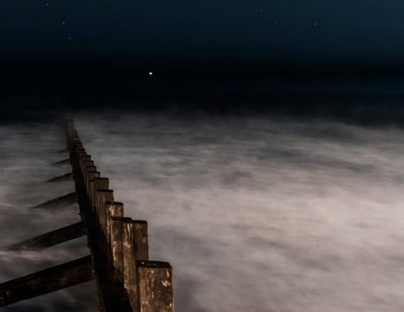 Een paalhoofd dat bij nacht de zee in loopt, onder een sterrenhemel