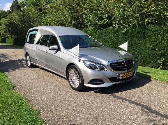 Een zilvergrijze rouwauto op een pad langs een groene haag