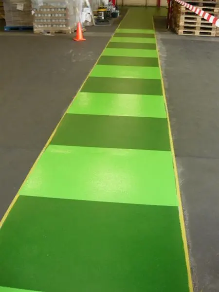 Een groen geschilderd looppad met lichtere dwarsbanen, dwars door een fabriekshal met pallets.