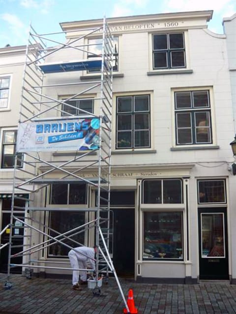 Rolsteiger met een banner van Baaijens tegen een monumentale gevel, met een schilder in witte werkkleding aan de voet.