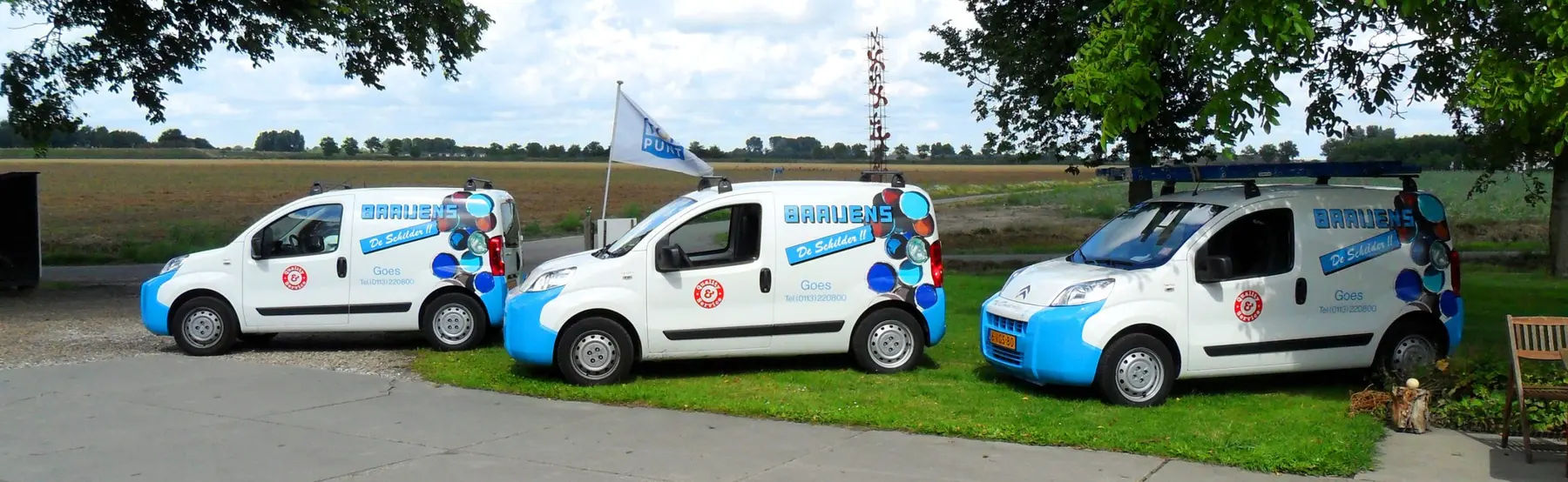 Drie wit-blauwe bestelbussen van Baaijens de Schilder naast elkaar aan de rand van een Zeeuws akkerland.