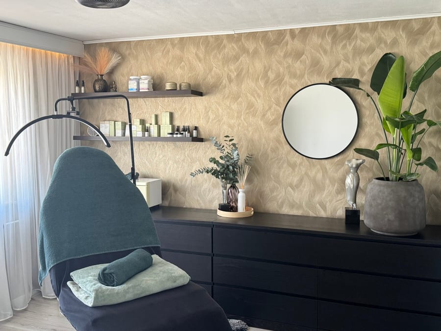 De behandelkamer: een donkerblauwe behandelstoel voor een wand met goudkleurig bladpatroon, een ronde spiegel, planten en een schap met producten.