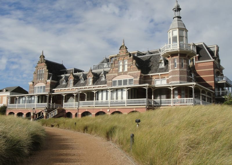 Monumentaal houten badpaviljoen met torentje, gezien vanaf het duinpad met helmgras ervoor.
