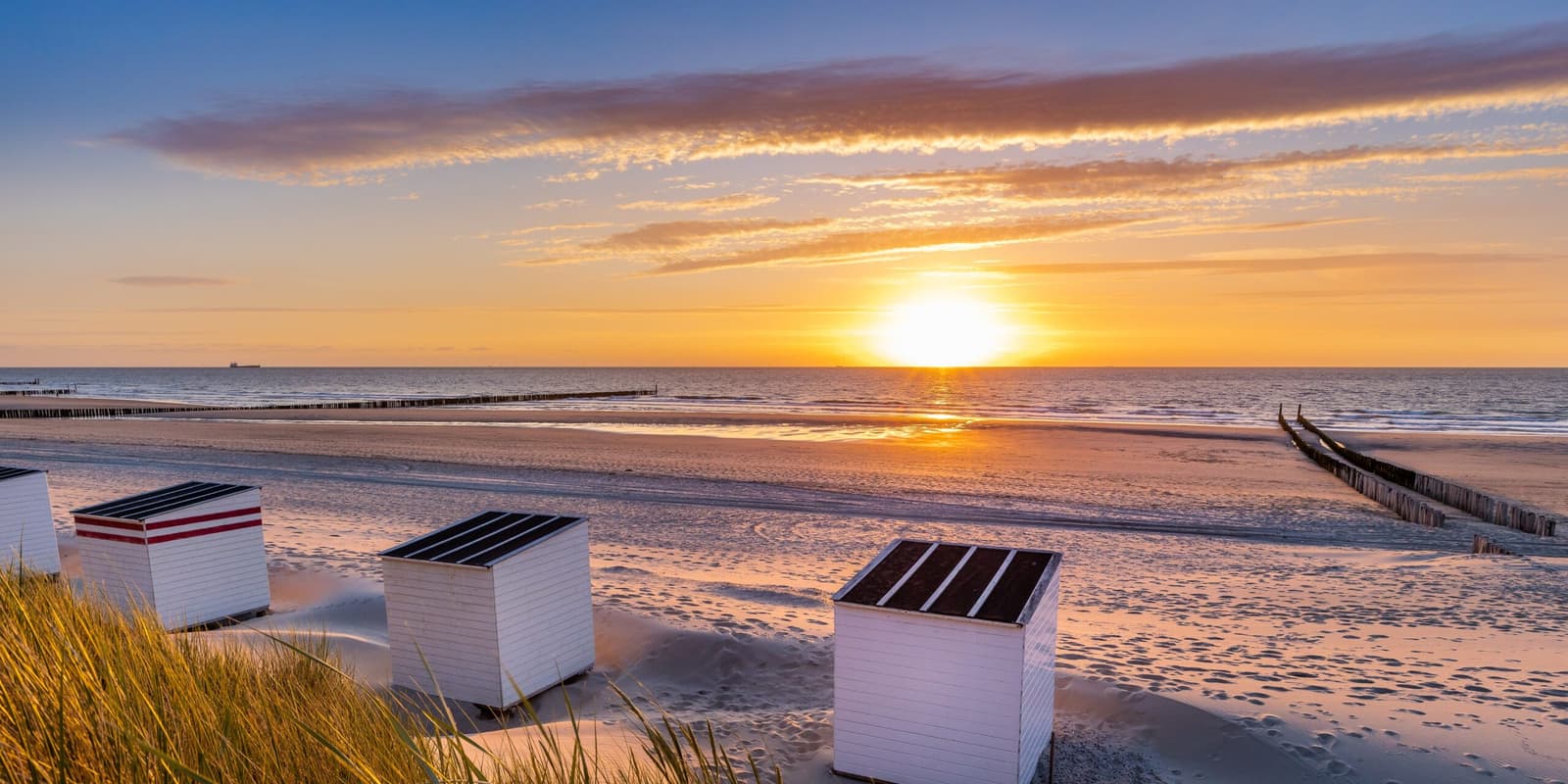 Strandcabines in het duingras bij Domburg, met de zon laag boven de zee en paalhoofden in het natte zand.