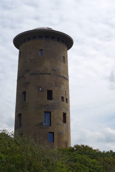 Ronde bakstenen watertoren boven op de duinrand, met struikgewas ervoor.