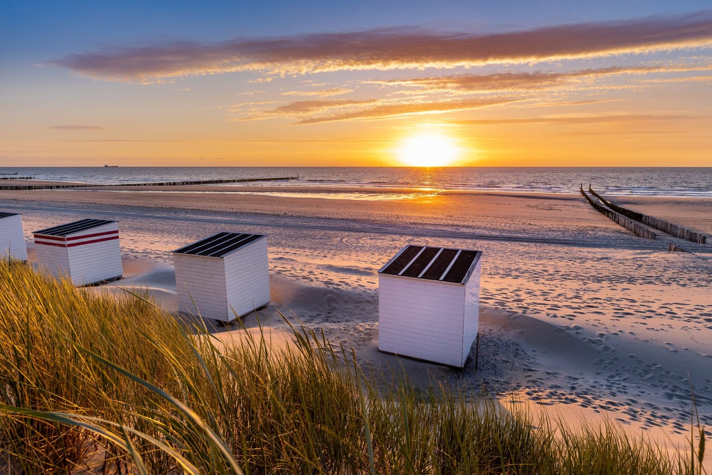 Twee witte strandcabines op het natte zand bij zonsondergang, met helmgras op de voorgrond.
