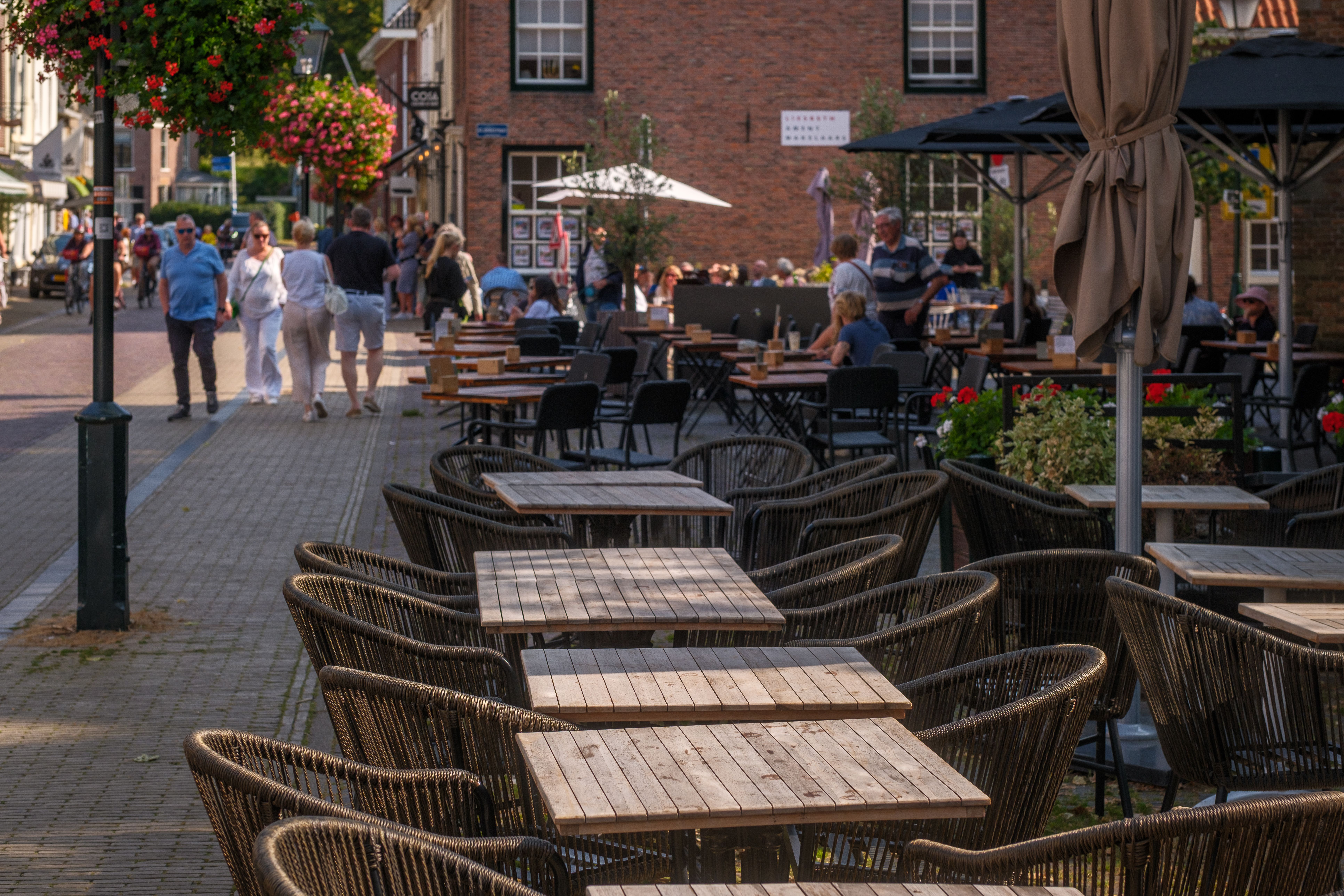 Terrasjes met rieten stoelen langs een dorpsstraat met bakstenen gevels en hangende bloembakken.