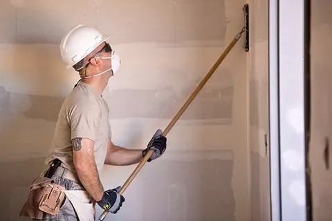 Schilder met helm en stofmasker bewerkt een wand met gereedschap op een verlengsteel, foto van de website van Elka Schildersbedrijf