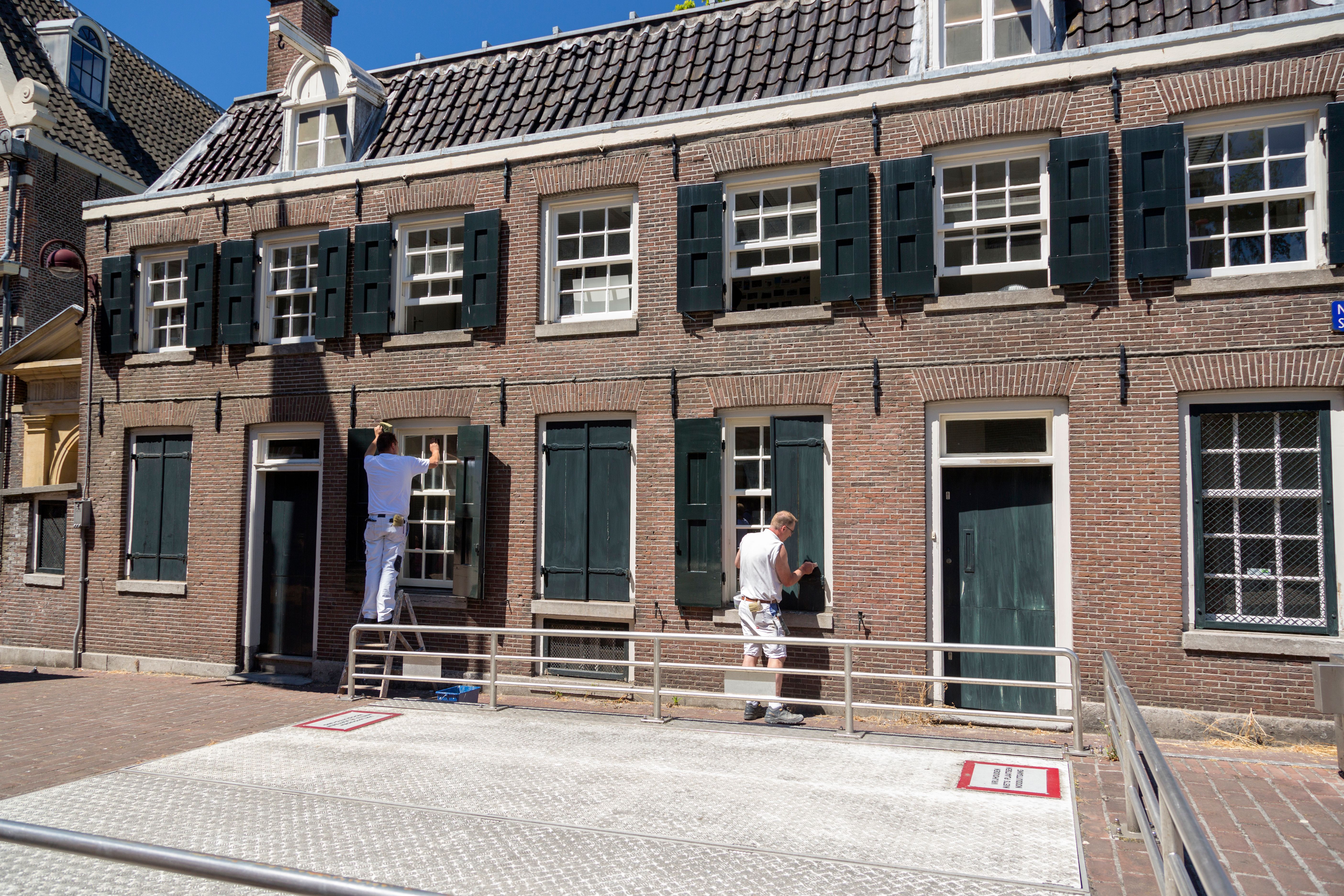 Twee schilders werken aan een bakstenen gevel met groene luiken, op een zonnige dag.