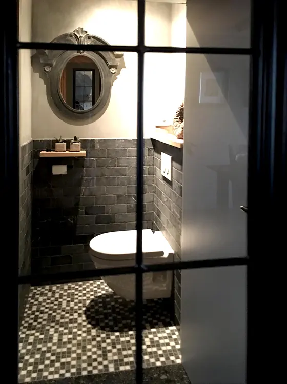 Zicht door een zwarte stalen deur op een toilet met donkere natuursteenstrips en een mozaïekvloer