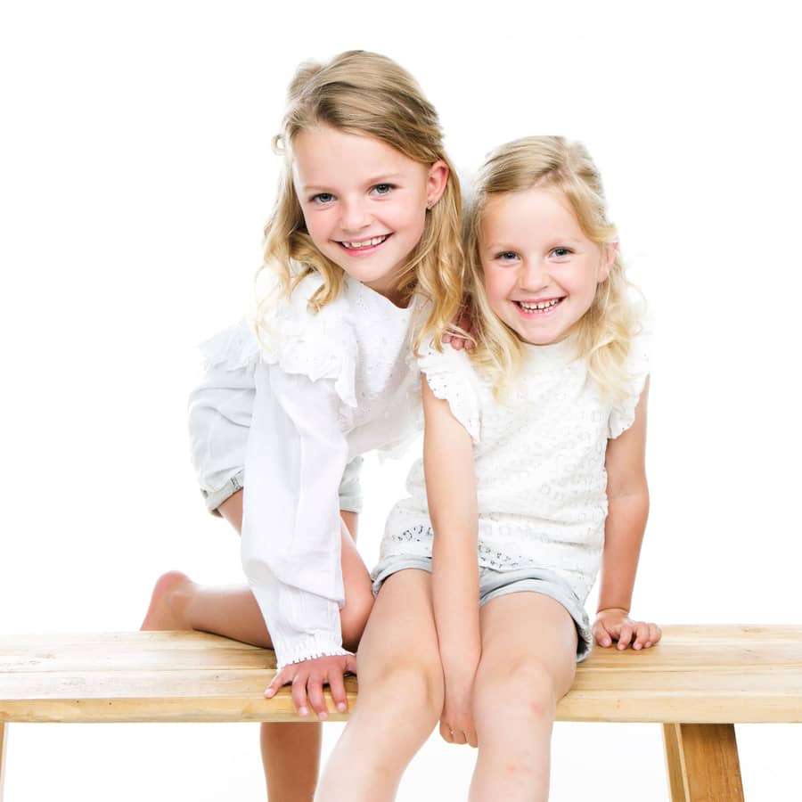 Twee zusjes in witte kleding lachen samen op een houten bankje