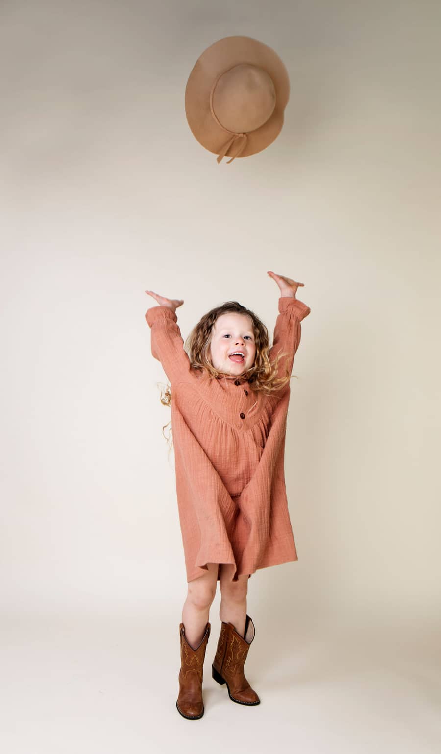 Meisje in een terracotta jurk met de armen omhoog tegen een zandkleurige achtergrond