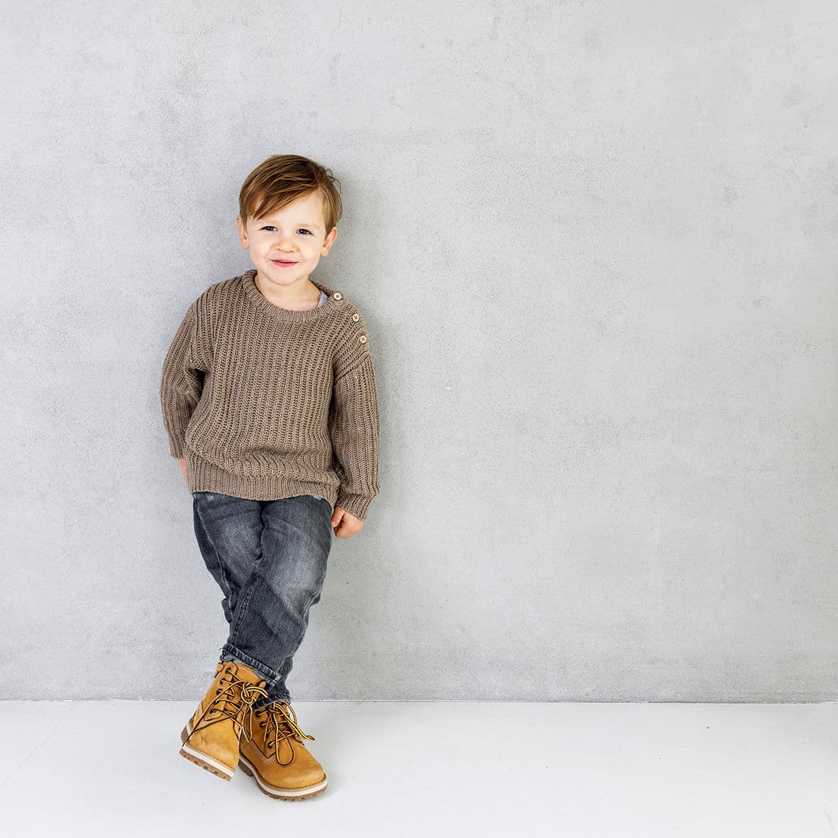 Jongen in een bruine trui tegen een lichtgrijze betonnen wand