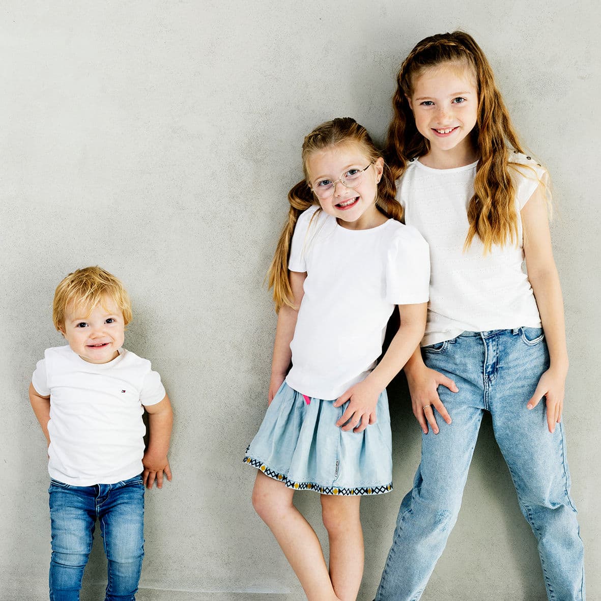 Drie kinderen in witte shirts en spijkerbroek tegen de betonnen wand van de fotostudio