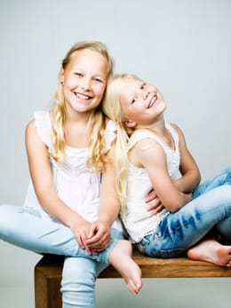Twee meisjes in witte topjes en jeans naast elkaar op een houten bankje, voor een licht grijze achtergrond.