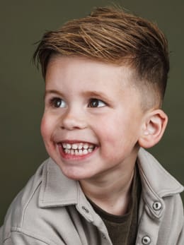 Lachende jongen in een beige jasje voor de olijfgroene achtergrond van de studio.