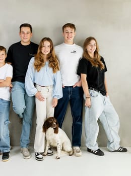 Groepsportret van zes personen met een hond, voor een lichte achtergrond.