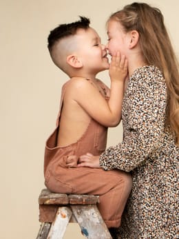 Jongen en meisje die elkaar bijna aanraken met hun neus, op een houten krukje voor het zandkleurige doek.