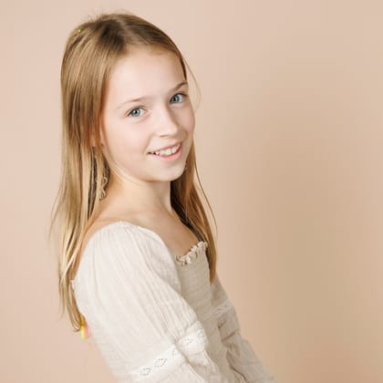 Meisje met lang blond haar in een witte kanten jurk, half weggedraaid voor een zandkleurige achtergrond.
