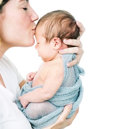 Moeder die haar pasgeboren baby, gewikkeld in een blauw gebreid doek, een kus op het hoofdje geeft.