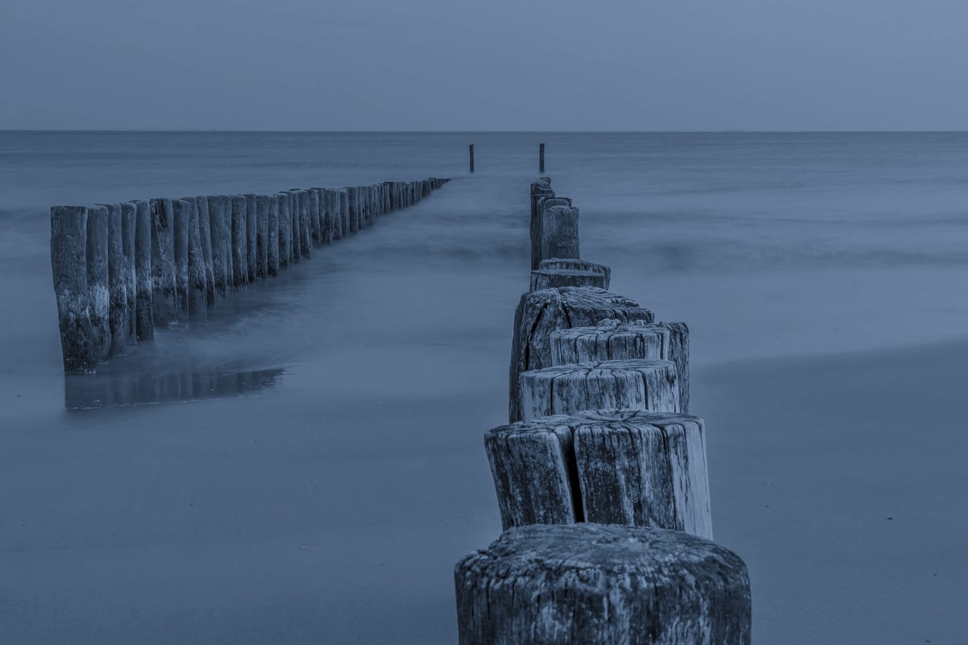 Twee rijen houten paalhoofden lopen bij schemer het vlakke water in, tot ze bij de horizon verdwijnen.