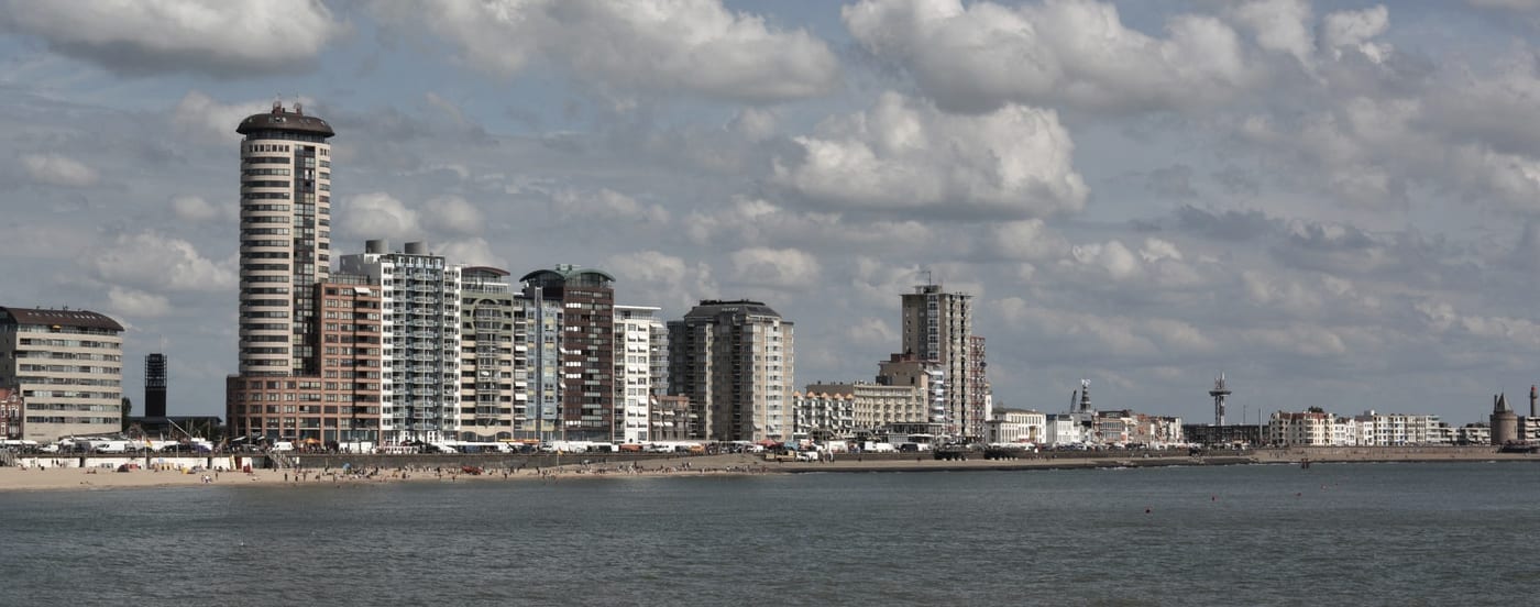 Een stad aan het water gezien van de overkant: woontorens en een strook strand onder een hoge wolkenlucht.