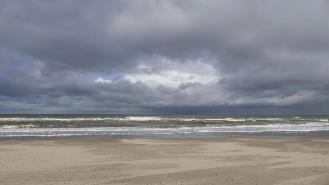 Een leeg, breed strand onder een zwaar bewolkte lucht, met de vloedlijn in het natte zand