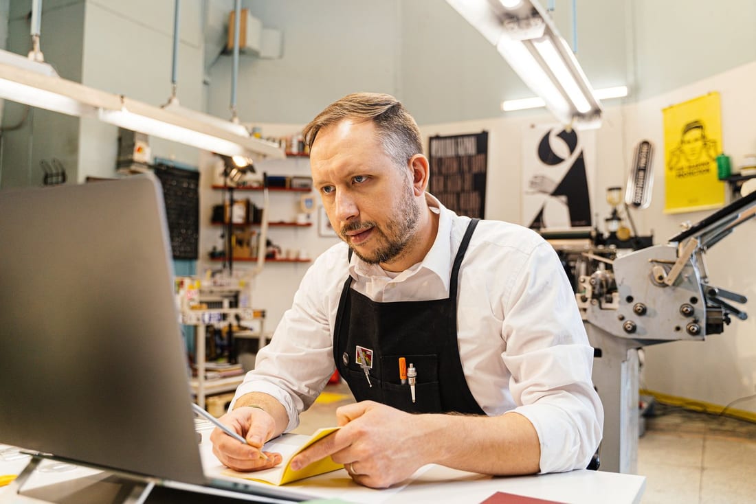 Iemand in een schort achter een laptop in een werkplaats, met een bedrukt vel papier in de hand.