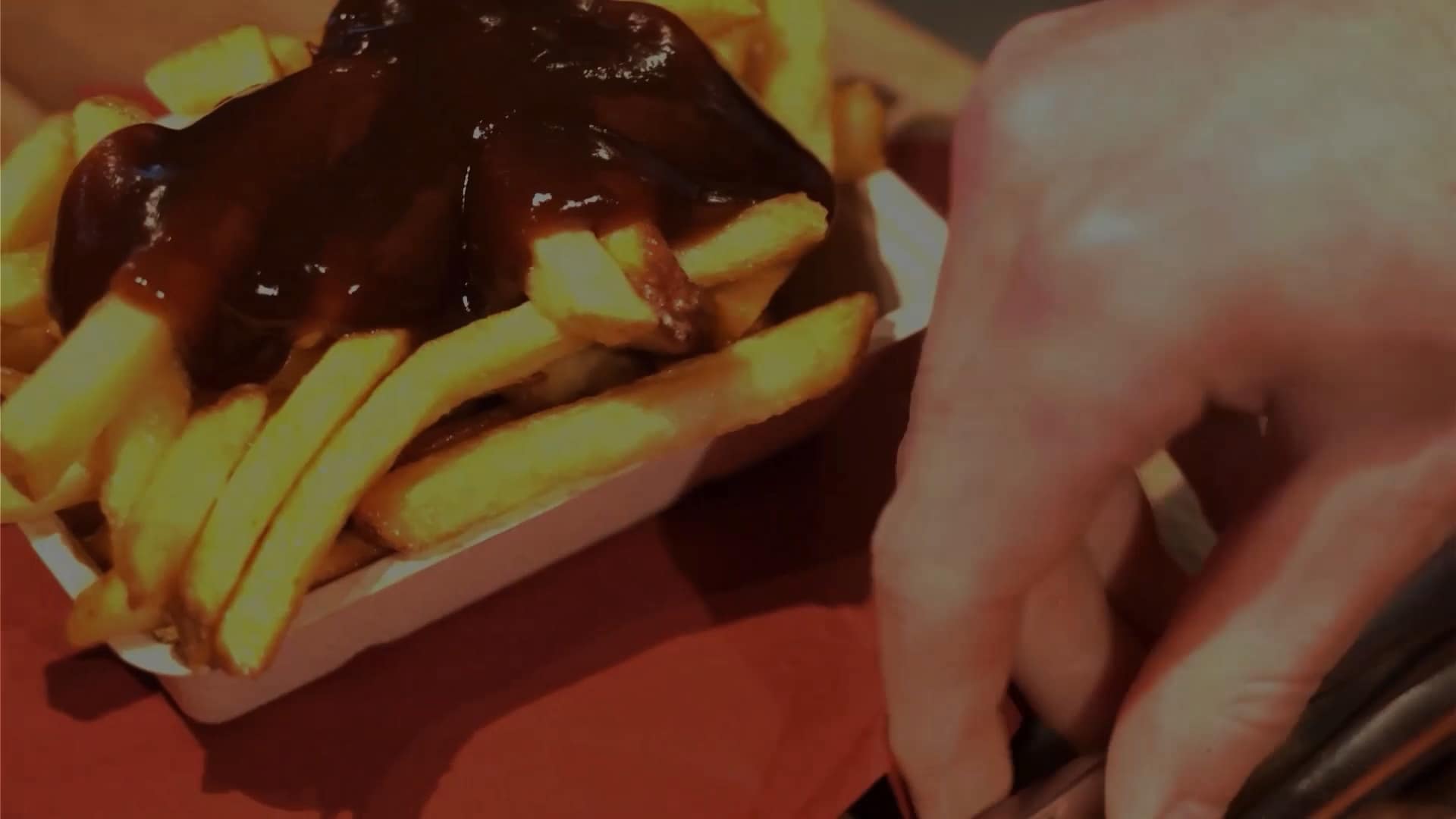 Bakje friet met donkere saus, een hand reikt ernaar in warm licht
