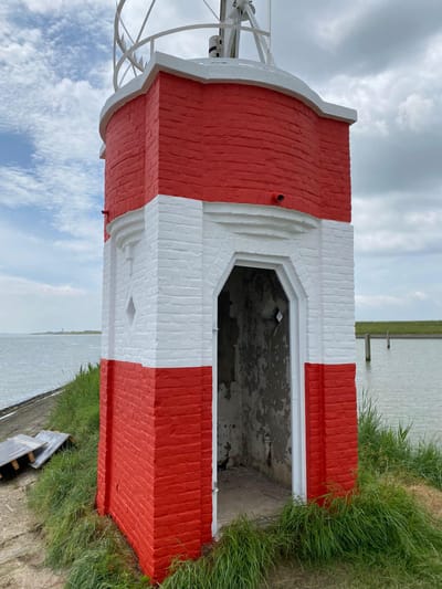 Kleine bakstenen toren op een dijk aan het water, geschilderd in brede banen rood en wit.