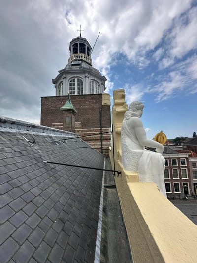Wit stenen beeld op een daklijst boven een leien dak, met de koepel van een kerktoren erachter.