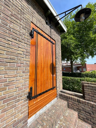 Houten luik met zwart smeedijzeren beslag in een bakstenen gevel, afgewerkt in een warme houtkleur.