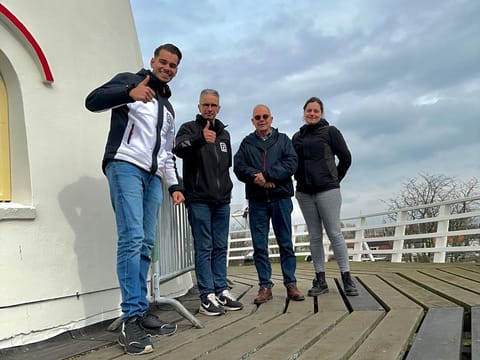 Vier collega&rsquo;s staan op een houten omloop tegen een witte molenromp, met bewolkte lucht erachter.