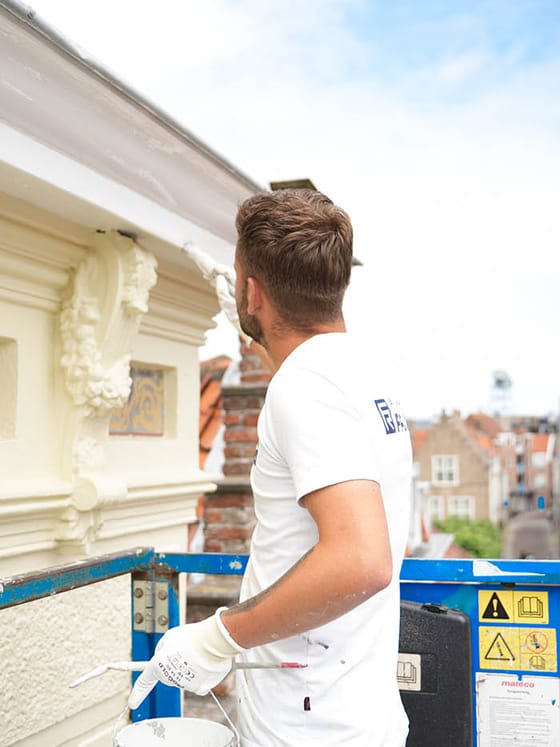 Schilder in bedrijfskleding op een hoogwerker naast een wit gestuukte gevel met ornamenten, met daken van de binnenstad op de achtergrond.