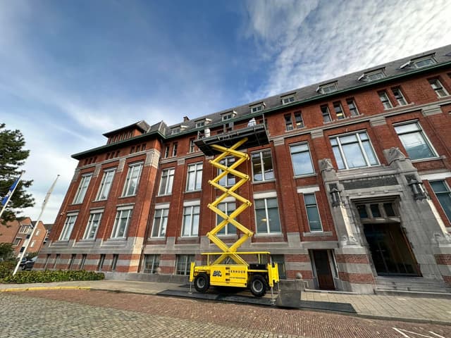 Twee schilders werken vanaf een schaarhoogwerker aan de daklijst van een grote bakstenen gevel.