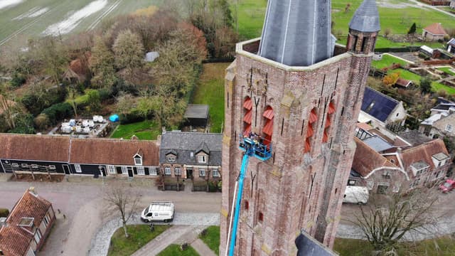 Een schilder werkt vanuit een hoogwerker aan de galmgaten van een bakstenen kerktoren.