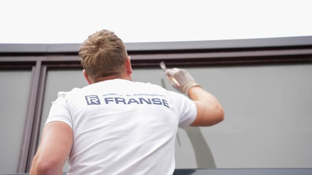 Een schilder in bedrijfskleding van Franse zet met een kwast een lijn op een grijs paneel.