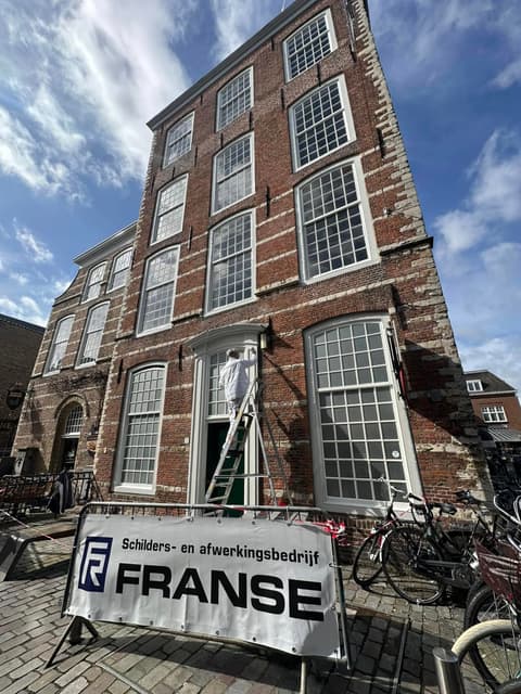 Een schilder op een ladder werkt boven de deur van een hoog historisch pand. Op het hek staat een spandoek van Franse.