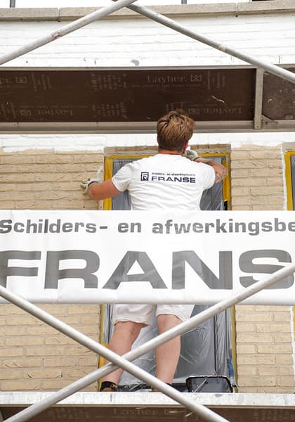 Een schilder van Franse werkt vanaf een steiger aan een kozijn in een lichte gevel. Aan de steiger hangt een spandoek van het bedrijf.