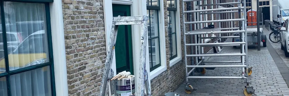 Trapladder, verfblik en een rolsteiger op straat voor een gemetselde gevel met witte kozijnen.