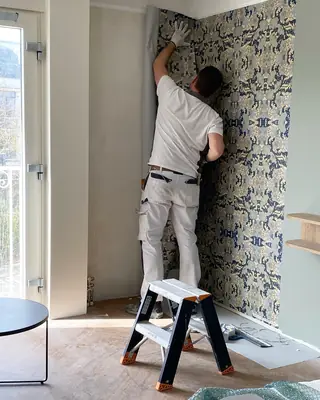 Schilder strijkt een baan blauw bloembehang glad, staand op een opstapje.