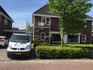 Witte bedrijfsbus voor een woning waar steigers tegen de gevel staan.