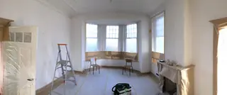 Leeggehaalde kamer met erker, kale wanden en een trapladder.