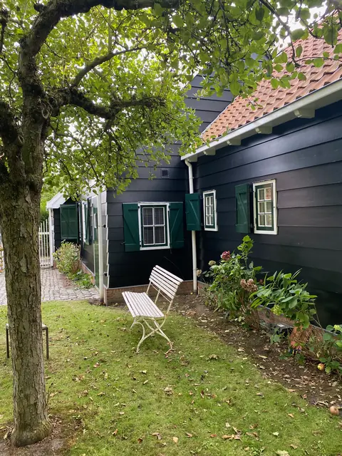Zwart geschilderd houten huis met witte kozijnen en donkergroene luiken, met een bankje onder een boom.